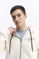 Campera con capucha y cierre frontal, color crema, con bolsillo canguro. Presenta un estampado de texto en el pecho y detalles en el puño.