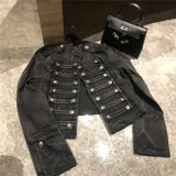 Chaqueta corta de denim negro con diseño de inspiración militar, cuello mao, cierre frontal con múltiples botones metálicos y detalles de trenzado horizontal. Presenta charreteras en los hombros y puños con botones decorativos.