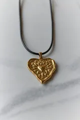Collar con dije de corazón en acero quirúrgico, con textura irregular y relieve, montado sobre un tiento encerado negro.