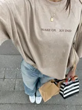 Remera oversize de color marrón topo con efecto acid wash, cuello redondo y manga corta. Cuenta con un pequeño estampado gráfico frontal con el texto 'WAKE UP IND' y una calavera.