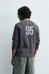 Camiseta de manga larga, corte ajustado (muscle fit), color gris oscuro con efecto lavado. Presenta un estampado frontal desgastado con el texto curvo "ST MONICA" en color plateado o gris claro.