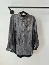 Camisa de corte holgado con acabado metalizado en tono plateado oscuro. Presenta cuello clásico, cierre frontal con botones, mangas largas con puños abotonados y un bolsillo frontal en el pecho.