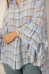 Camisa de manga larga con diseño a cuadros en tonos celestes, marrones y blancos. Presenta corte asimétrico con largo irregular, más larga en la parte trasera, y mangas amplias con volados escalonados.