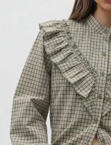 Camisa de manga larga con estampado de cuadros vichy en tonos verde oliva y crudo. Presenta cuello mao, cierre frontal con botones y volados decorativos en la parte delantera. Las mangas terminan en puños elastizados con un ligero acabado de volado.
