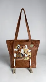 Cartera tipo tote de cuero color marrón terra, con diseño pintado a mano en el frente y borlas decorativas colgantes.