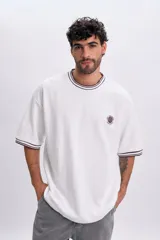 Remera blanca de manga corta con ribetes a rayas marrones en cuello y puños, corte oversize y pequeño bordado circular en el pecho.