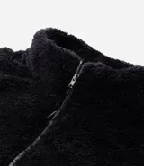 Buzo de tejido peludo color negro, con cuello alto y cierre frontal parcial. Presenta mangas largas con puños elásticos, bolsillo tipo canguro y etiqueta decorativa en el pecho.
