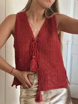 Chaleco de punto calado color rojo con destellos metalizados, escote en V profundo y cierre frontal mediante cordones tejidos con borlas decorativas. Presenta un corte recto y holgado.
