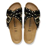 Sandalias Birkenstock modelo Siena Rivet, color negro, con diseño de tiras cruzadas de gamuza decoradas con tachas doradas y una hebilla metálica grande ajustable. Presentan una plantilla anatómica de corcho y una suela estriada.