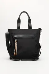 Bolso tipo tote color negro, con doble asa superior y correa desmontable. Presenta un bolsillo frontal con solapa, bolsillos laterales y un detalle colgante decorativo con tiras de colores.