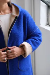 Tapado de paño color azul con capucha, cierre frontal y detalles en color beige en el interior de la capucha, puños y bordes de los bolsillos.