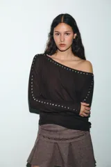 Camiseta de cuello barco y manga larga tipo murciélago, confeccionada en tejido de punto gris melange. Presenta detalle de tachas metálicas a lo largo de los hombros y las mangas.