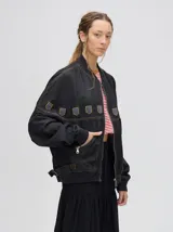 Campera tipo bomber confeccionada en denim de algodón color negro. Presenta un diseño holgado con cierre frontal metálico, bolsillos laterales con cierre y detalles decorativos en forma de escudos en la parte superior delantera.