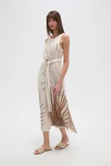 Vestido largo sin mangas, color crudo, con estampado de hojas en tonos marrones y negros. Presenta un cuello redondo y un lazo de tela a juego que ajusta la cintura. La falda es fluida y tiene aberturas laterales.