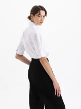 Camisa de popelina blanca con bordados de pequeñas bocas que forman un patrón de rayas. Presenta un diseño de corte holgado, mangas cortas con volumen, cuello clásico con broche metálico y cartera oculta con botones al tono.