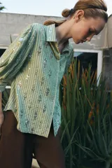 Camisa de corte recto en color verde claro con estampado de rayas verticales y detalles de lentejuelas brillantes. Presenta cuello solapa, cierre frontal con botones y mangas tres cuartos.