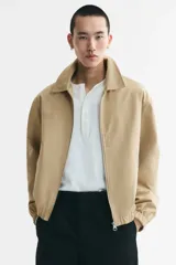 Campera tipo bomber de sarga de algodón en color beige, con cuello inglés, cierre frontal metálico y bolsillos diagonales ribeteados. Presenta terminaciones con elástico en los puños y el ruedo.