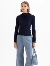 Sweater de tejido de punto fino color marrón, con ajuste al cuerpo, cuello alto con detalle de volados y moño, y mangas largas con puños acampanados con volados.