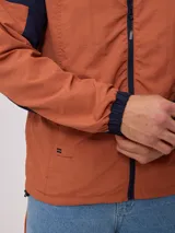 Campera rompevientos con cierre frontal, confeccionada en nylon con diseño de bloques de color en tonos verde azulado y gris claro. Cuenta con capucha, puños elásticos y cordón de ajuste en el ruedo.