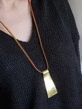 Collar con dije rectangular irregular de alpaca color dorado, colgando de una tira de cuero marrón.