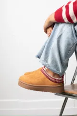 Zueco de gamuza color tostado con interior forrado en piel sintética. Presenta una banda decorativa bordada con motivos étnicos en el borde superior y una suela de plataforma plana de goma.