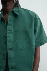 Camisa de denim de algodón color verde oscuro, de corte relajado (relaxed fit) y manga corta. Posee cuello solapa, bolsillo plastrón en el pecho con bordado a tono y cierre frontal de botones.