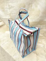 Bolso tipo tote grande con estampado a rayas verticales en tonos celeste, blanco y bordó. Tiene doble asa corta de mano y cierre superior.