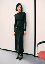 Vestido largo de punto color gris oscuro, con cuello perkins y manga larga. Presenta un diseño entallado con detalle fruncido en la cintura y una anilla metálica decorativa.