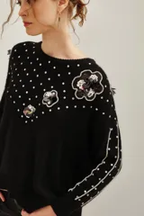 Sweater negro de punto con cuello redondo, manga larga y diseño holgado. Presenta aplicaciones de lentejuelas plateadas dispersas, bordes plateados en las mangas y apliques florales con lentejuelas en el hombro y frente.