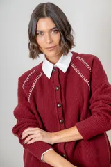 Saco de punto color beige, con cierre frontal de botones marrones y detalle de costuras decorativas oscuras en los hombros. Presenta un diseño clásico con cuello redondo.