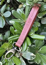 Llavero de cuero en formato de tira con diseño minimalista, confeccionado en cuero genuino marrón con interior en contraste color fucsia, pespuntes rosados y herrajes metálicos envejecidos con gancho tipo mosquetón.