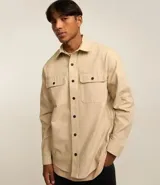 Camisa de manga larga para hombre confeccionada en sarga de algodón color beige. Presenta un diseño con cuello inglés, cierre frontal de botones oscuros y dos bolsillos con solapa en el pecho.
