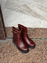Bota de caña media color marrón rojizo, con diseño de cuero liso, cierre lateral y detalle de correa con hebilla en la parte superior. Cuenta con suela track gruesa de goma negra.