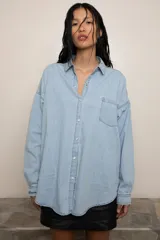 Camisa de jean liviano en tono celeste claro, con corte oversize, cuello clásico, cierre frontal con botones metálicos, bolsillo en el pecho y detalle de bordado en el puño.