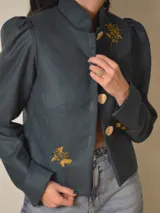 Chaqueta de terciopelo azul marino con hombreras marcadas, mangas largas y bordados florales plateados en el frente. Presenta un diseño estructurado con cierre frontal de botones.