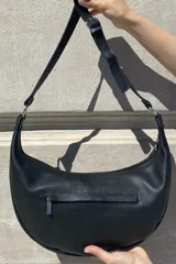 Cartera bandolera de cuero negro con diseño de media luna, decorada con tachas metálicas plateadas. Cuenta con correa regulable, cierre principal y bolsillo interior con forro.