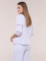 Blusa blanca de calce suelto y caída ligera, con escote en V y cuello Mao pequeño. Presenta mangas tres cuartos abullonadas con elástico en los puños y detalles de calado o puntilla encastrada. Espalda lisa con canesú y leve frunce.
