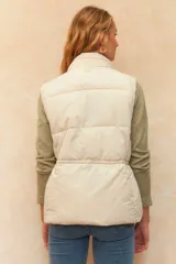 Chaleco puffer color crudo, sin mangas, con cuello alto, cierre frontal con cremallera, bolsillos frontales con solapa y cordón ajustable en la cintura.