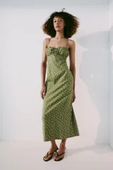 Vestido midi verde militar con estampado de lunares rosados, de corte recto y tirantes finos cruzados en la espalda. Presenta escote con frunces y detalle de lazo en el centro.