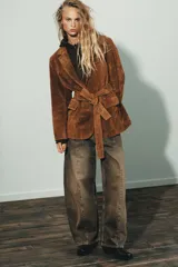 Blazer de gamuza color marrón con cuello solapa, manga larga con hombreras y cierre frontal con botones. Incluye bolsillos con solapa en el delantero, bolsillo de vivo en el pecho y cinturón de lazada en el mismo material.
