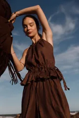 Blusa color marrón oscuro, estilo halter con cuello fruncido y pechera con elástico ajustable en la cintura. Presenta un volante con borde deshilachado y tiras trenzadas regulables que se cruzan en la espalda.