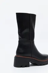 Bota de caña media en cuero sintético color negro, con diseño minimalista y suela robusta dentada con detalle de borde en color marrón.