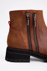 Botin de cuero vacuno color suela, con diseño de caña media, doble cierre lateral y suela track negra de goma.