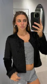 Chaqueta corta de textura peluda en color negro, con cuello redondo y cierre frontal abierto. Presenta mangas largas y un corte recto.