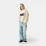 Sweater de punto color crema con finas rayas horizontales en beige. Presenta un logo grande con la palabra "EME" en azul marino en el frente y tiene cuello redondo y corte holgado.