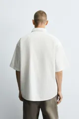 Camisa de algodón blanca con finas rayas verticales negras, corte relajado (relaxed fit) y manga corta. Presenta un bordado en hilo rojo en el lado izquierdo del pecho.
