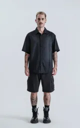 Camisa de manga corta en color negro con proceso de lavado, cuello clásico y cierre frontal con botones. Presenta un diseño minimalista con una costura vertical decorativa en el frente.