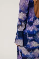 Vestido largo con estampado abstracto en tonos azul, lila y blanco. Tiene cuello mao con abertura en forma de lágrima, mangas largas acampanadas, espalda abierta, tajo frontal y flecos en el ruedo.