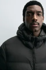 Campera puffer negra con capucha ajustable con elásticos, cuello alto, cierre frontal con cremallera, bolsillos laterales y puños elásticos ajustables.