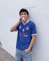 Camiseta de fútbol de manga corta en color azul con diseño estampado abstracto y cuello en V rosado con detalles blancos a juego con los puños. Presenta inscripciones y escudos deportivos en el pecho.